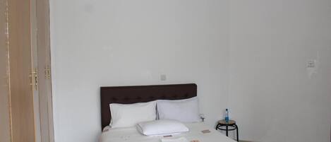 A neatly made bed with white linens and a brown headboard, a small bedside table with a water bottle, and a closed wooden wardrobe.