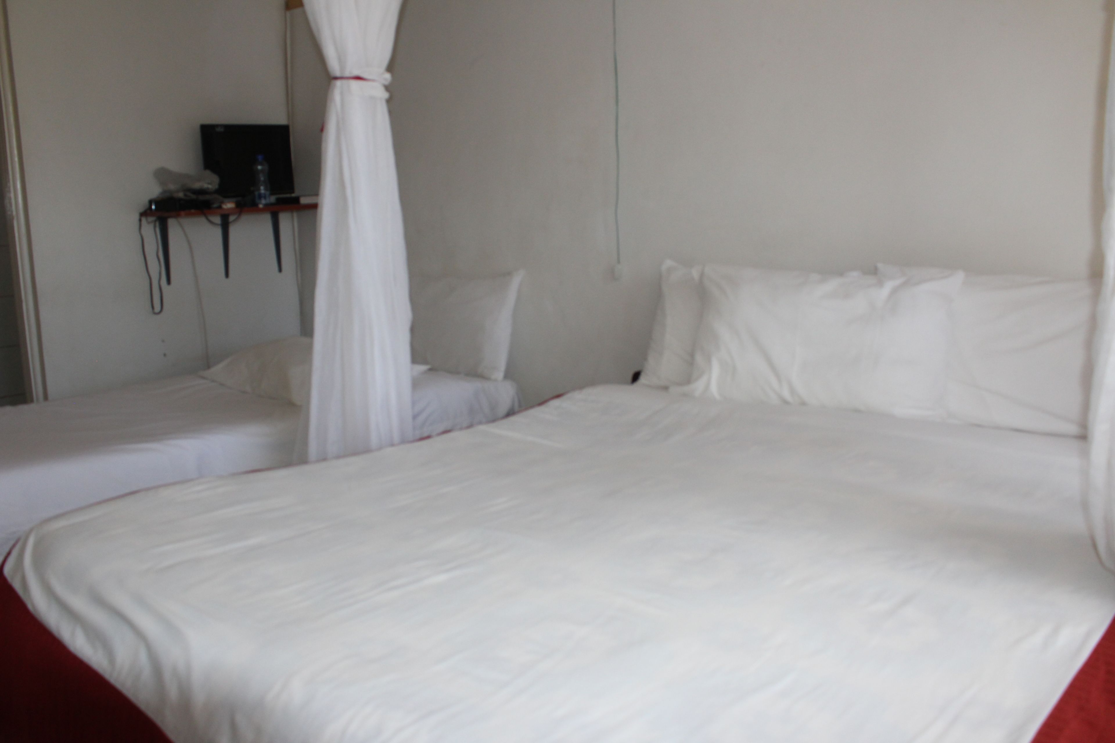 Une chambre avec deux lits simples, chacun doté de draps blancs et surmonté d’un ciel de lit. On y trouve une petite table sur laquelle sont posés un ordinateur portable et une bouteille d’eau.