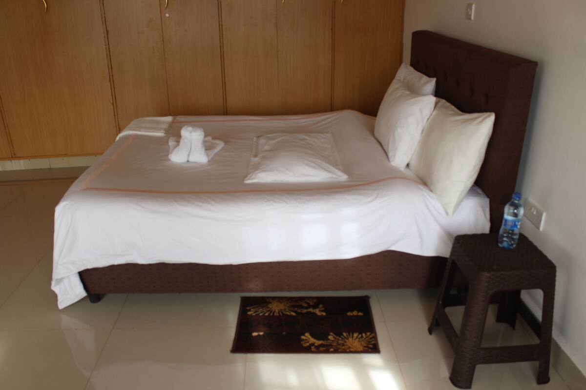Una cama bien hecha con sábanas y almohadones blancos, un cabecero de madera, un taburete pequeño de madera con una botella, y una alfombra decorativa en el suelo.