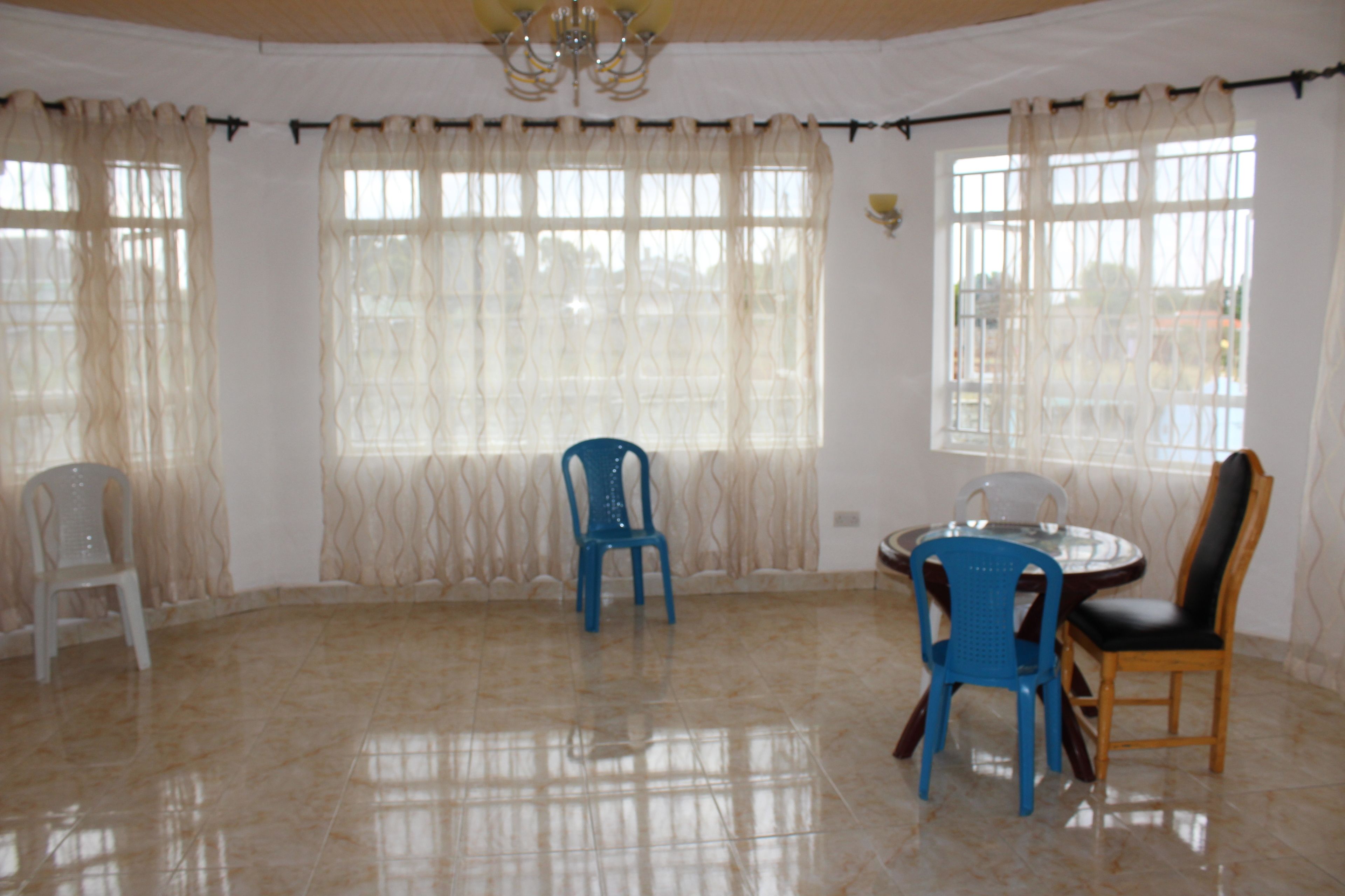Una habitación con piso de baldosas, una mesa redonda con sillas y ventanas grandes con cortinas.