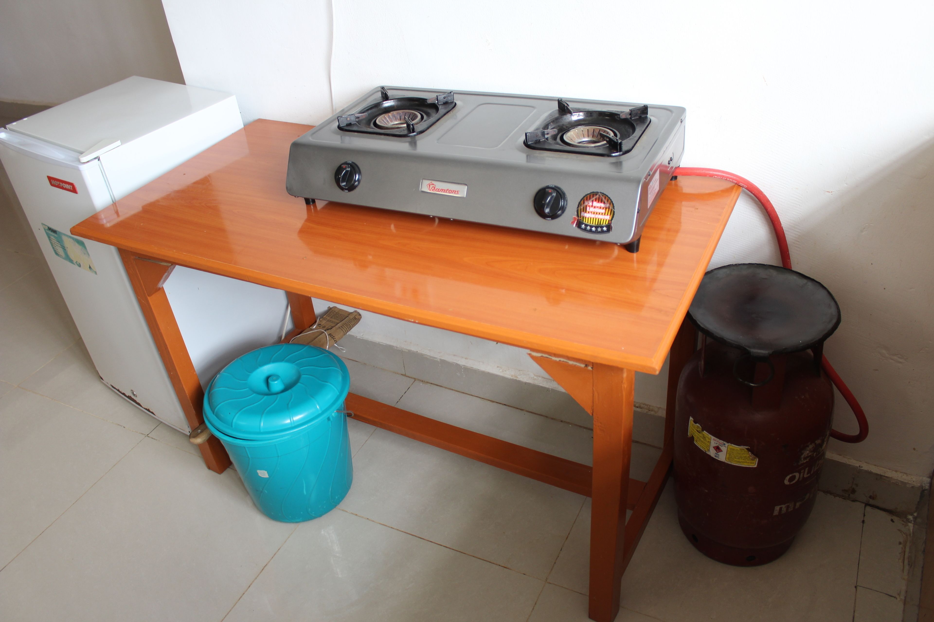 A compact kitchen with a gas stove, a small refrigerator, a blue trash bin, and a propane tank.