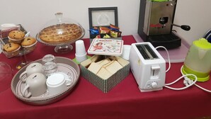 A table with a red tablecloth, a coffee maker, a toaster, a tray with teacups and cookies, a framed picture, and a bowl of muffins.