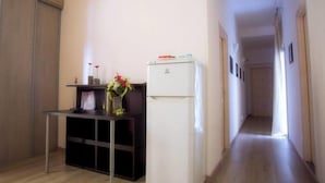A compact kitchen with a white refrigerator, a small table with a vase, and a dark cabinet. A hallway extends from the kitchen.