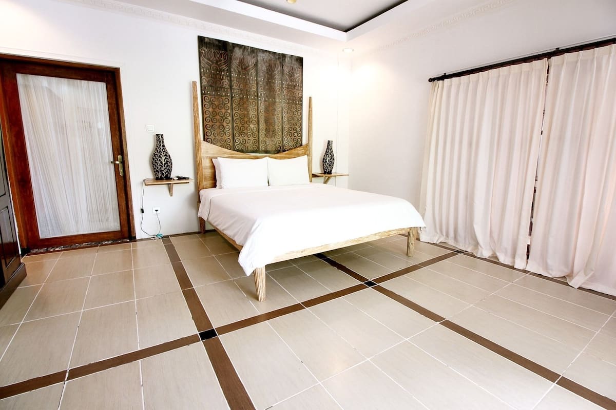 A hotel room with a bed, a wooden headboard, a large tapestry, a curtain, and a tiled floor.