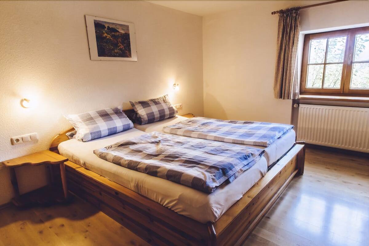Ein Doppelbett mit kariertem Bettbezug, zwei Nachttische, eine Wandleuchte, ein Fenster mit Vorhängen und eine Heizung.