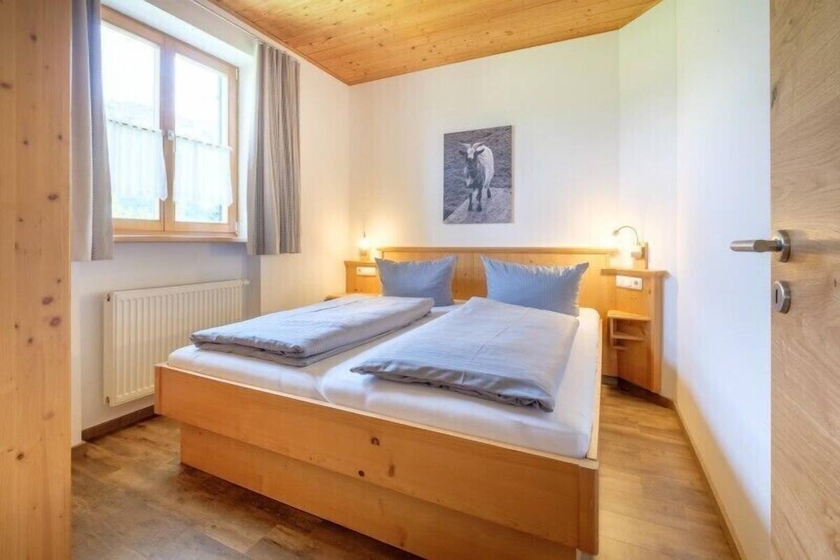 Ein Schlafzimmer mit einem hölzernen Bett, zwei Nachttischen, einem Schranken und einem Fenster mit Vorhängen, sowie einem an der Wand befestigten Bild.