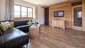 A modern living room with a wooden floor, a black leather sofa with green cushions, a wooden coffee table, and a TV mounted on a wooden wall. - Haflingerhof - Kematsried (Bad Hindelang)