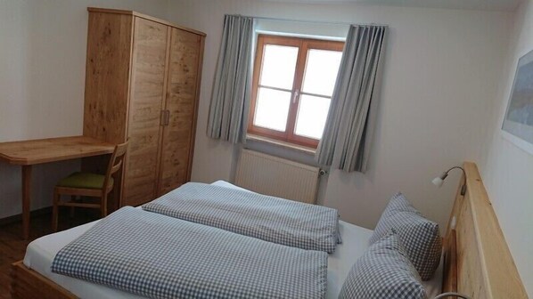 A bedroom with a wooden wardrobe, a desk, a chair, a bed with checkered bedding, and a window with curtains.