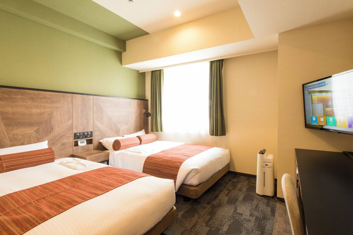 A hotel room with two beds, a desk, a chair, a TV, and a window with curtains.