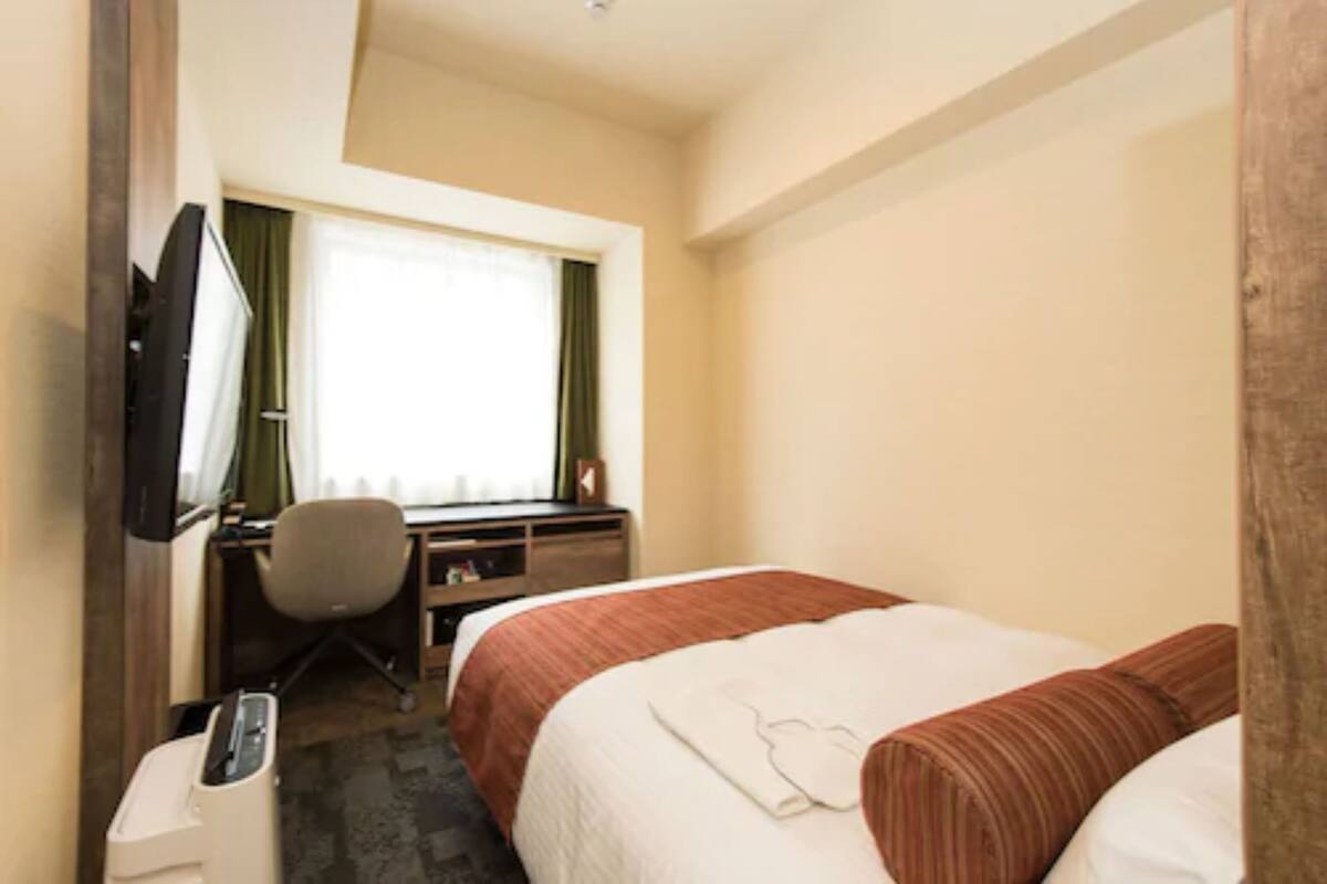 A hotel room with a bed, a desk with a chair, a television, and a window with curtains.