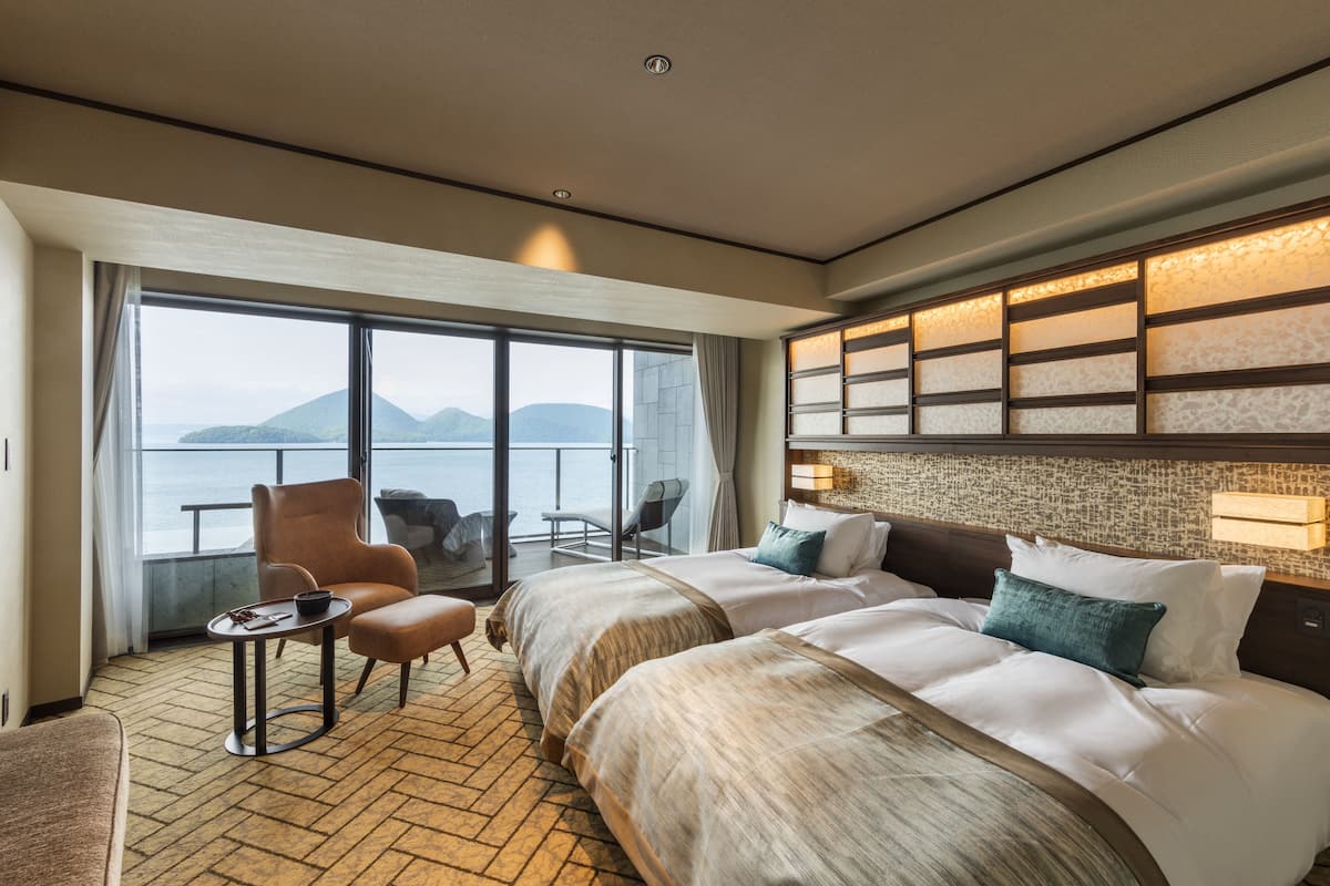 A hotel room with a large window, a balcony, a bed with white linens, and a seating area with a table and chairs.