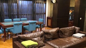 A living room with a brown leather sofa, a glass-top dining table with blue chairs, and a large window with dark curtains. - Beo-River Chocolate (Belgrade)