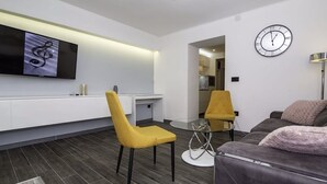 A modern living room with a sofa, two yellow chairs, a glass coffee table, a flat-screen TV mounted on the wall, and a clock.