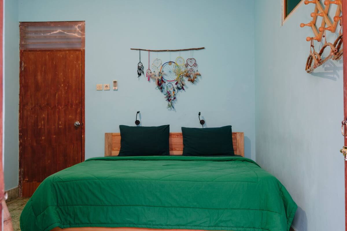 Ein Schlafzimmer mit Holztür, einem Bett mit grüner Bettdecke, zwei Kissen und einem Traumfänger an der Wand.