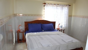 A bedroom with a wooden bed, two blue pillows, a white bedspread, a nightstand with a lamp, and a window with sheer curtains. - Chambre en Ville (Sen Monorom)