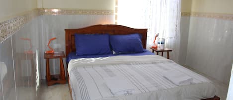 A bedroom with a wooden bed, two blue pillows, a white bedspread, a nightstand with a lamp, and a window with sheer curtains.
