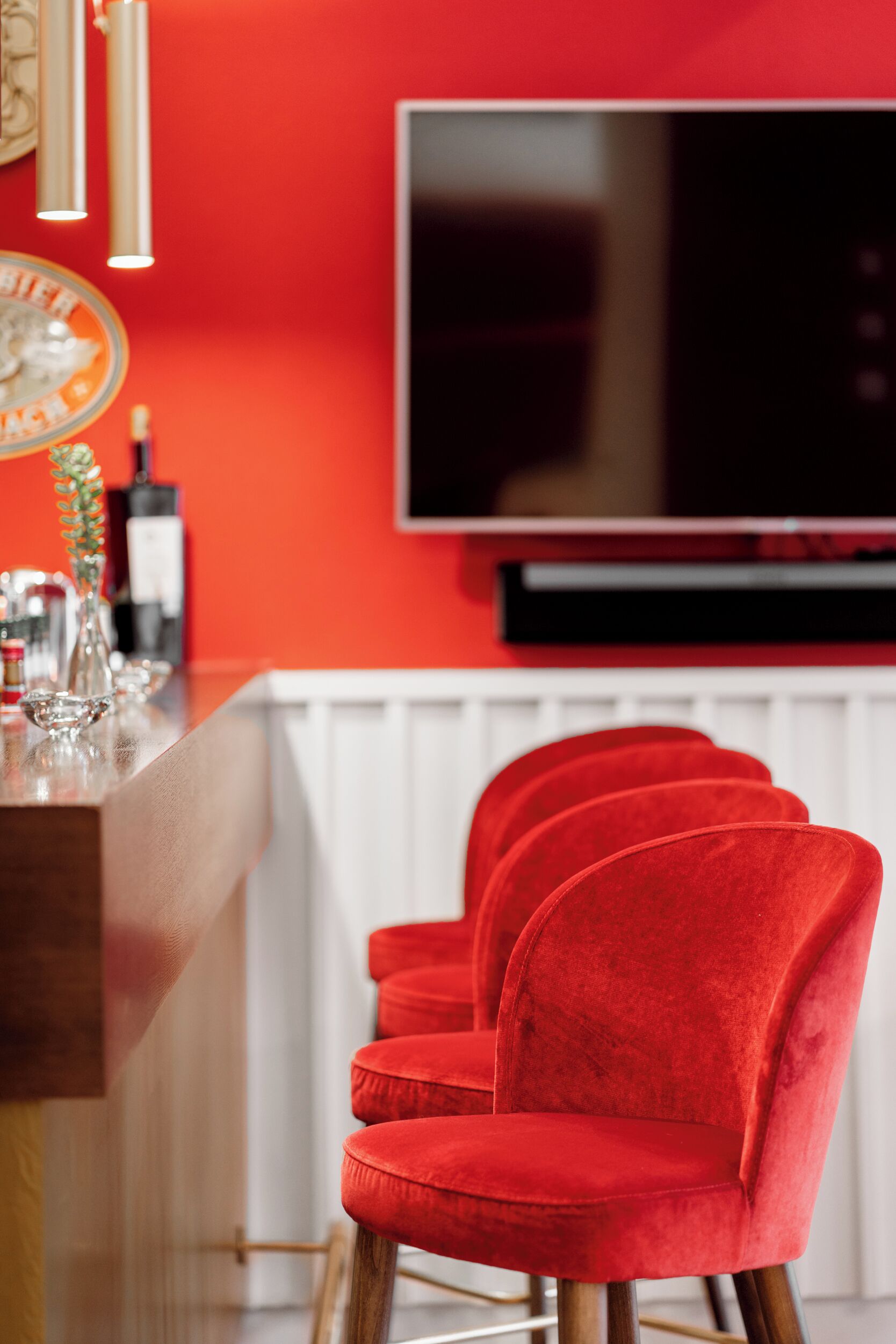 Una zona de bar con una pared roja, un televisor de pantalla plana y tres taburetes de terciopelo rojo.
