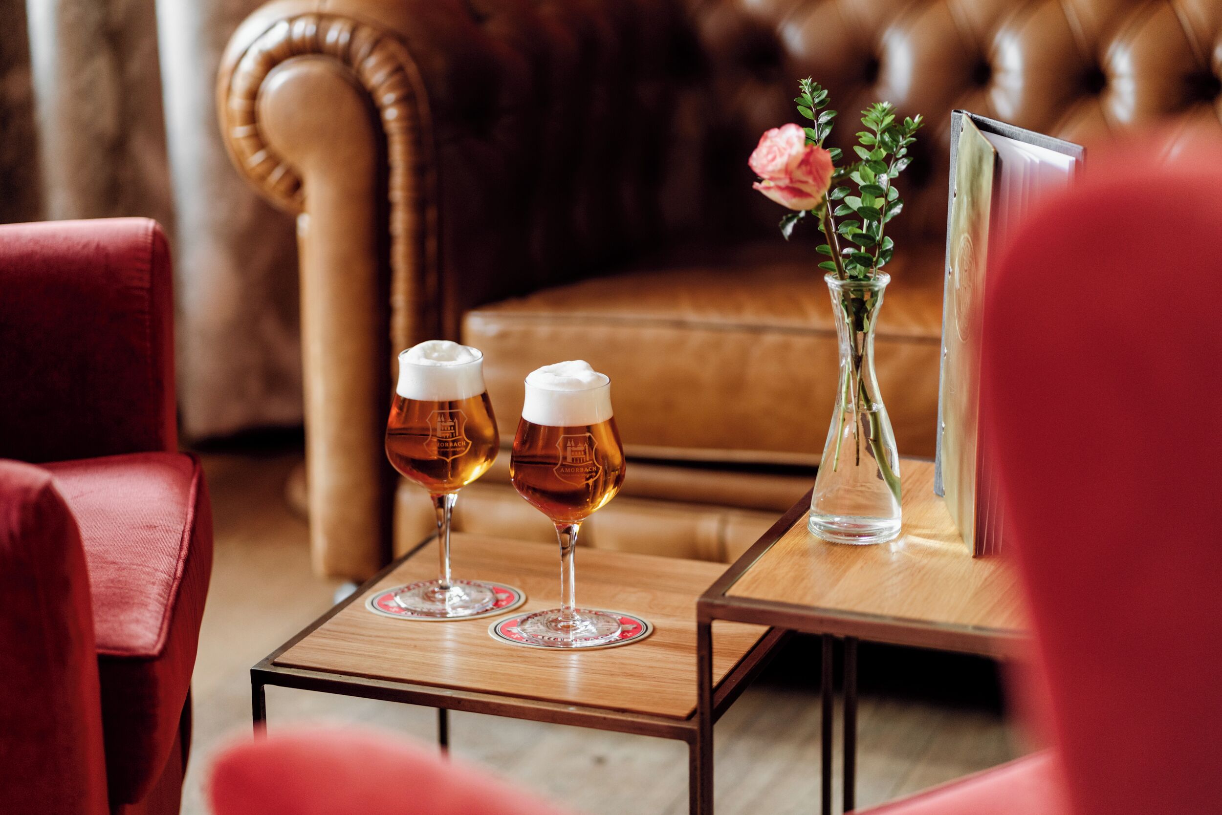 Two glasses of beer on a wooden table with a vase of flowers and a framed picture.