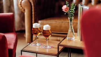 Two glasses of beer on a wooden table with a vase of flowers and a framed picture.