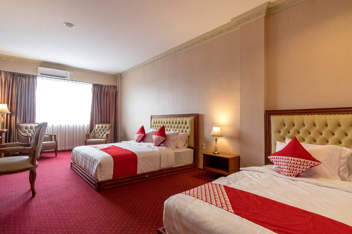 A hotel room with two beds, a sitting area with chairs, a window with curtains, and an air conditioning unit.