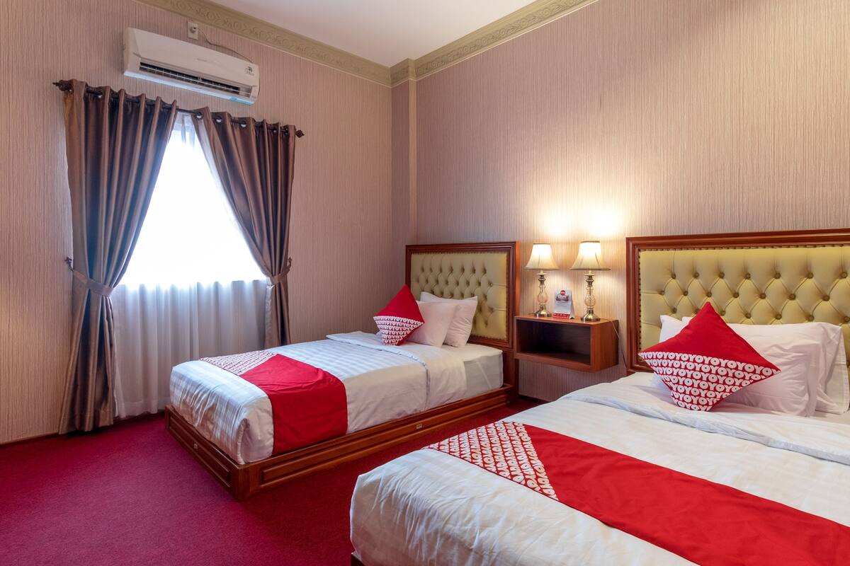 A hotel room with two beds, red and white bedding, a large window with curtains, an air conditioning unit, and bedside lamps.