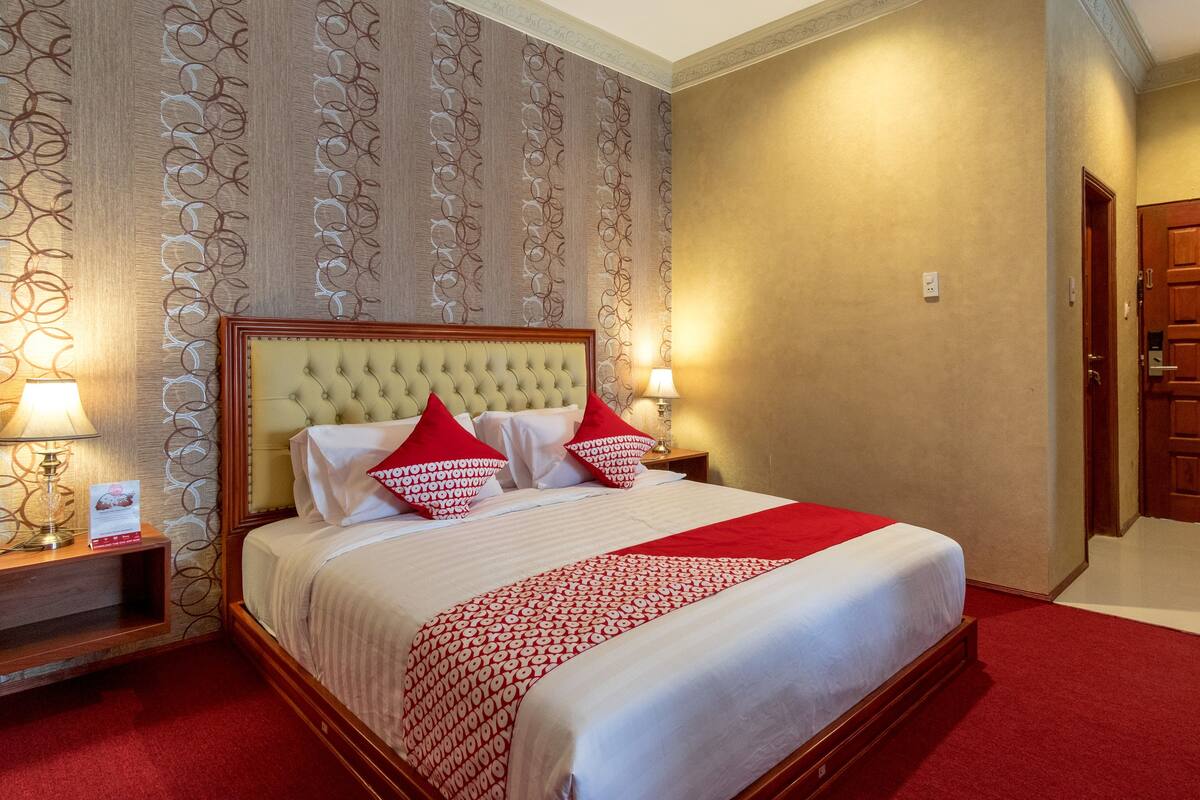A neatly made bed with a red and white bedspread, two red pillows, and a wooden headboard. There are two bedside lamps on wooden nightstands. The room has a patterned wallpaper and a plain beige wall.
