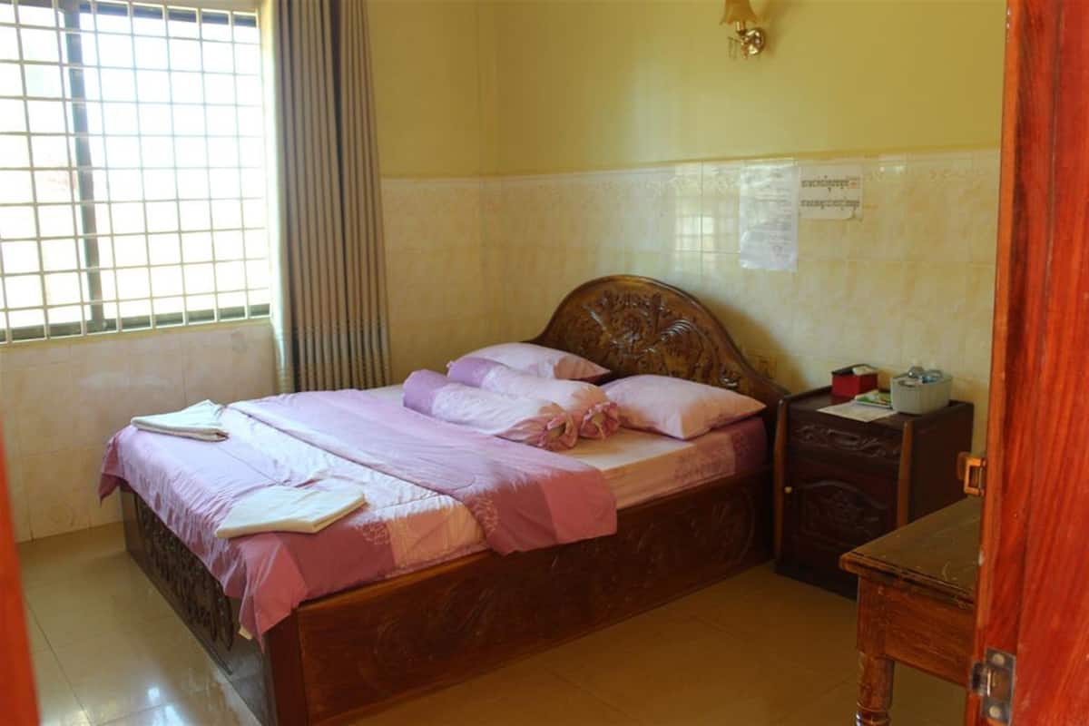 Ein ordentlich bezogenes Bett mit rosa Bettwäsche, ein Holz-Kopfteil, ein Nachttisch mit Lampe und ein Fenster mit Jalousien.