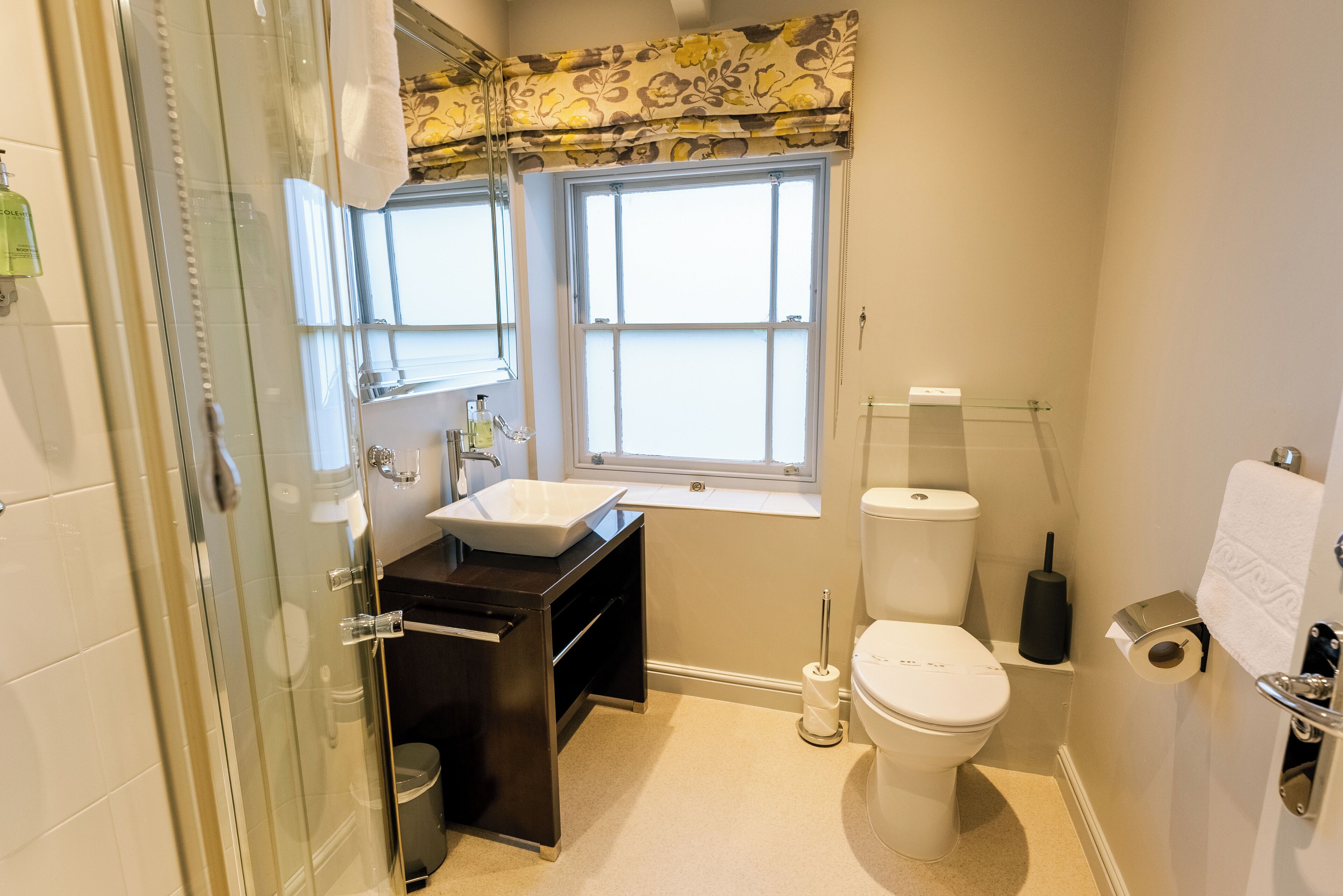 A bathroom with a glass shower enclosure, a white sink, a toilet, and a window with a floral curtain.