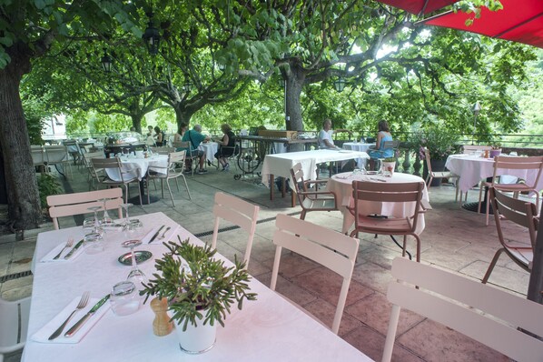An outdoor dining area with tables and chairs under large trees, surrounded by greenery. - Hotel Beau Site Notre Dame (Rocamadour)