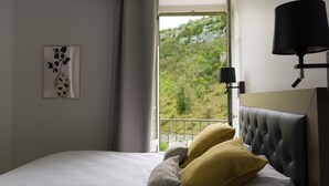 Un dormitorio con cama, ventana con vista a un bosque, lámpara de noche y un cuadro en la pared.