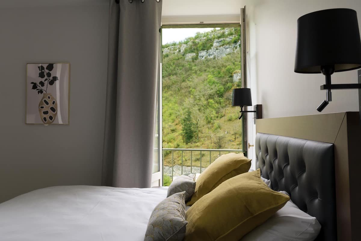 Een slaapkamer met een bed, een raam met uitzicht op een bos, een bedlamp en een schilderij aan de muur.