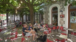 Un café al aire libre con mesas y sillas bajo un gran árbol, rodeado de muros de piedra y un arco de piedra.