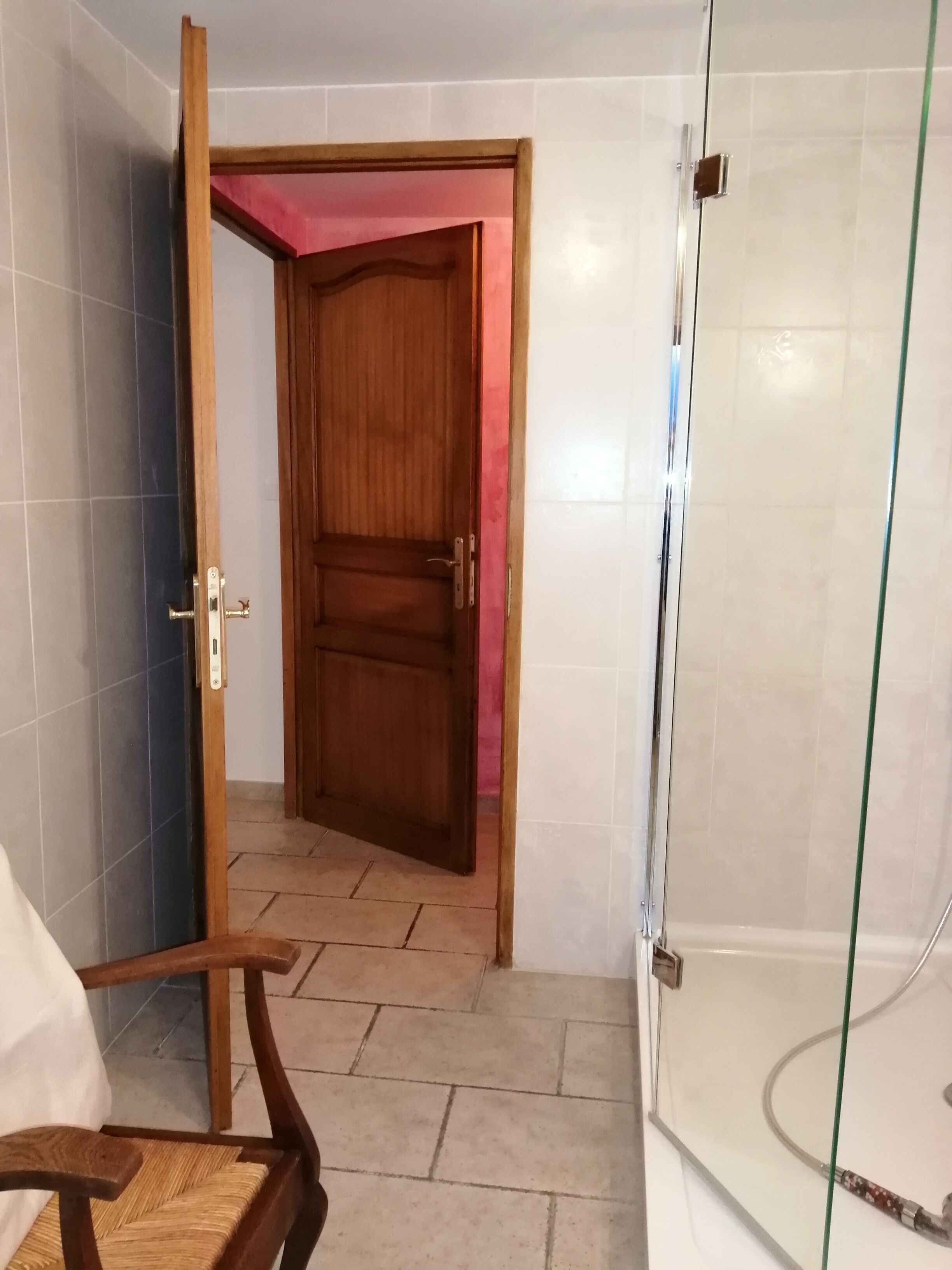 Ein Zimmer mit Holztür, Fliesenwänden, einem gefliesten Boden und einer Duschkabine aus Glas, sowie einem Stuhl.
