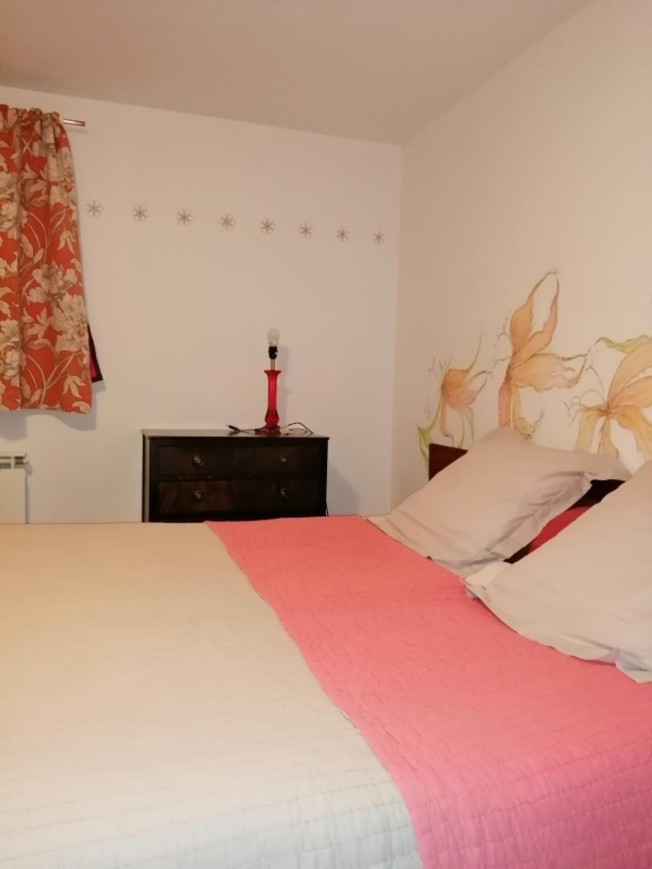 A bedroom with a bed, a nightstand, a lamp, and a wall with floral artwork.