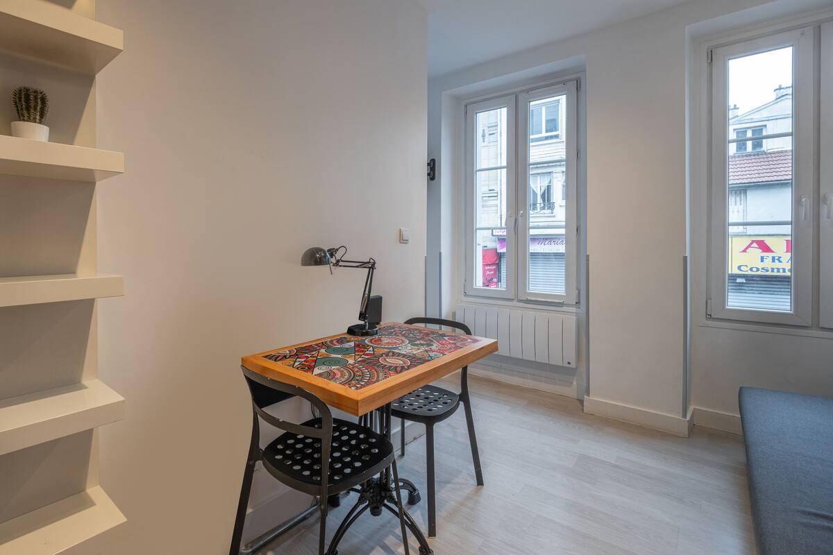 A small room with a wooden table, two chairs, a white shelf with a cactus, and a window with a view of a building.