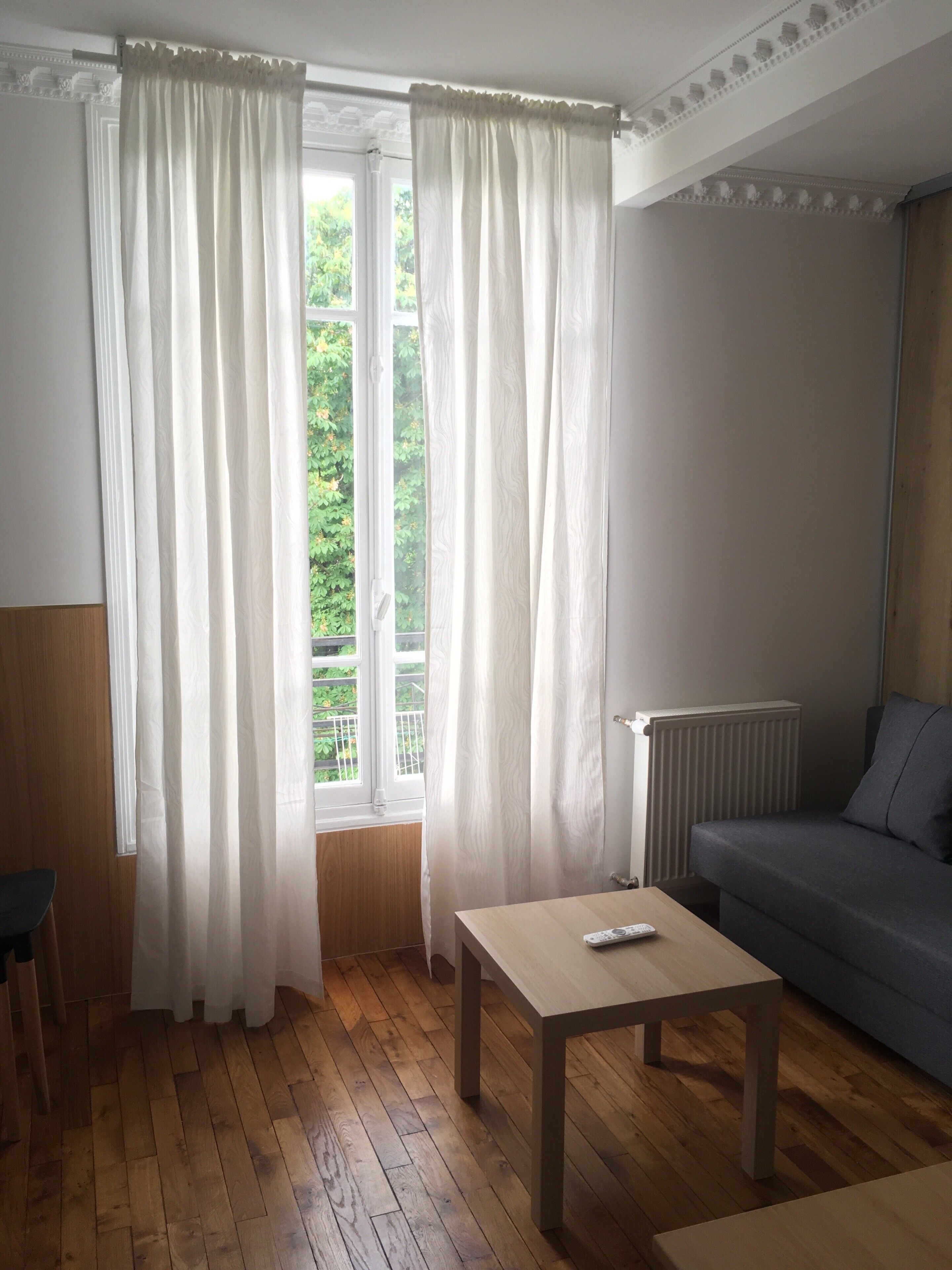 A room with wooden flooring, a sofa, a table with a remote control, and a window with white curtains.