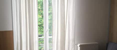 A room with wooden flooring, a sofa, a table with a remote control, and a window with white curtains.