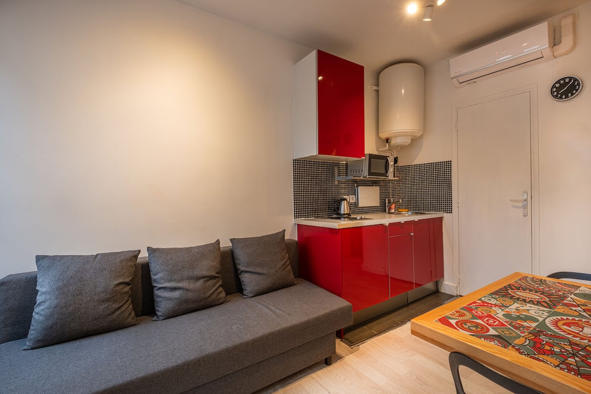 A compact living space with a grey sofa, a red kitchen area, and a dining table with a patterned tablecloth.