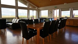A spacious room with a wooden table and chairs, large windows offering a view of the countryside, and a sofa area. - Guesthouse Uggi (Rangárþing ytra)