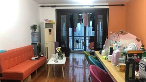 A compact living space with a sofa, a small table with a vase, a chair, a desk with various items, and a view of a cityscape through a glass door. - Pinkhostelling laternational (Shanghai)