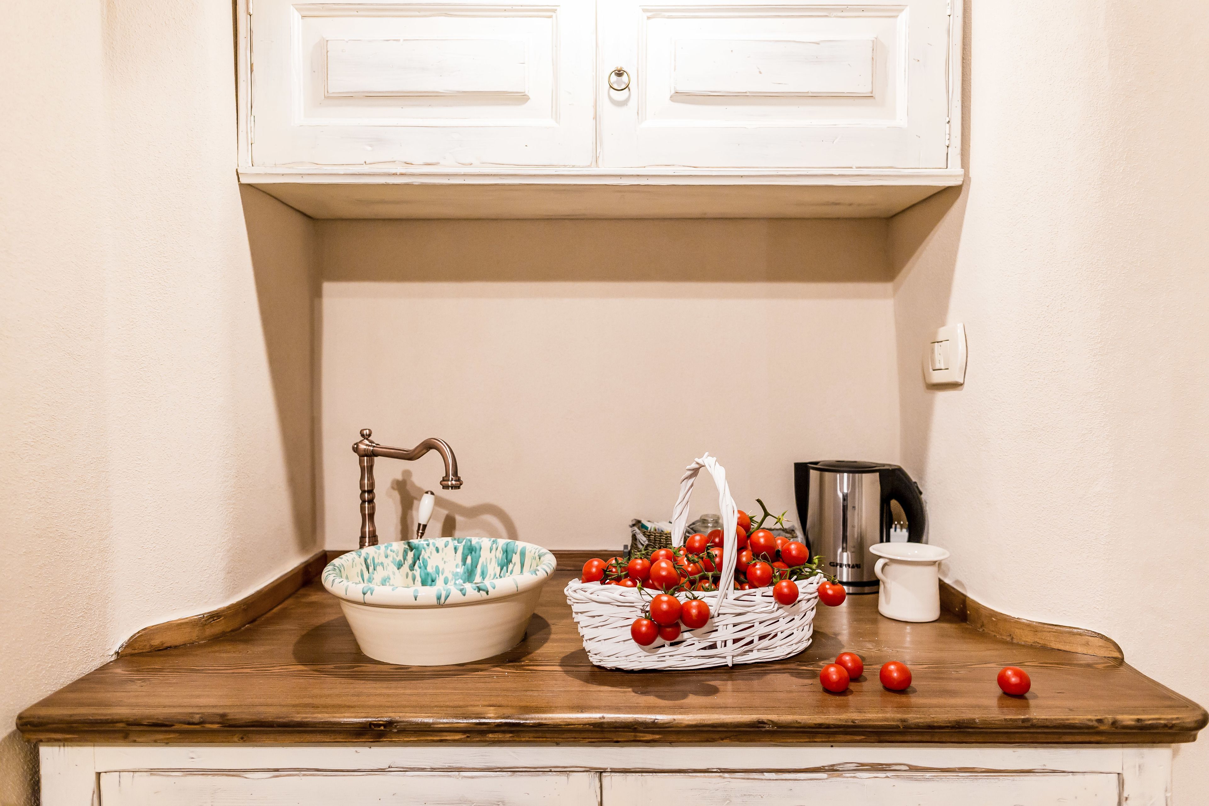 Una encimera de cocina con un lavamanos blanco, una cesta de tomates, un hervidor y una jarra blanca.