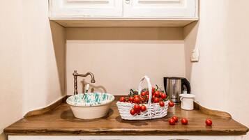 Una encimera de cocina con un lavamanos blanco, una cesta de tomates, un hervidor y una jarra blanca.