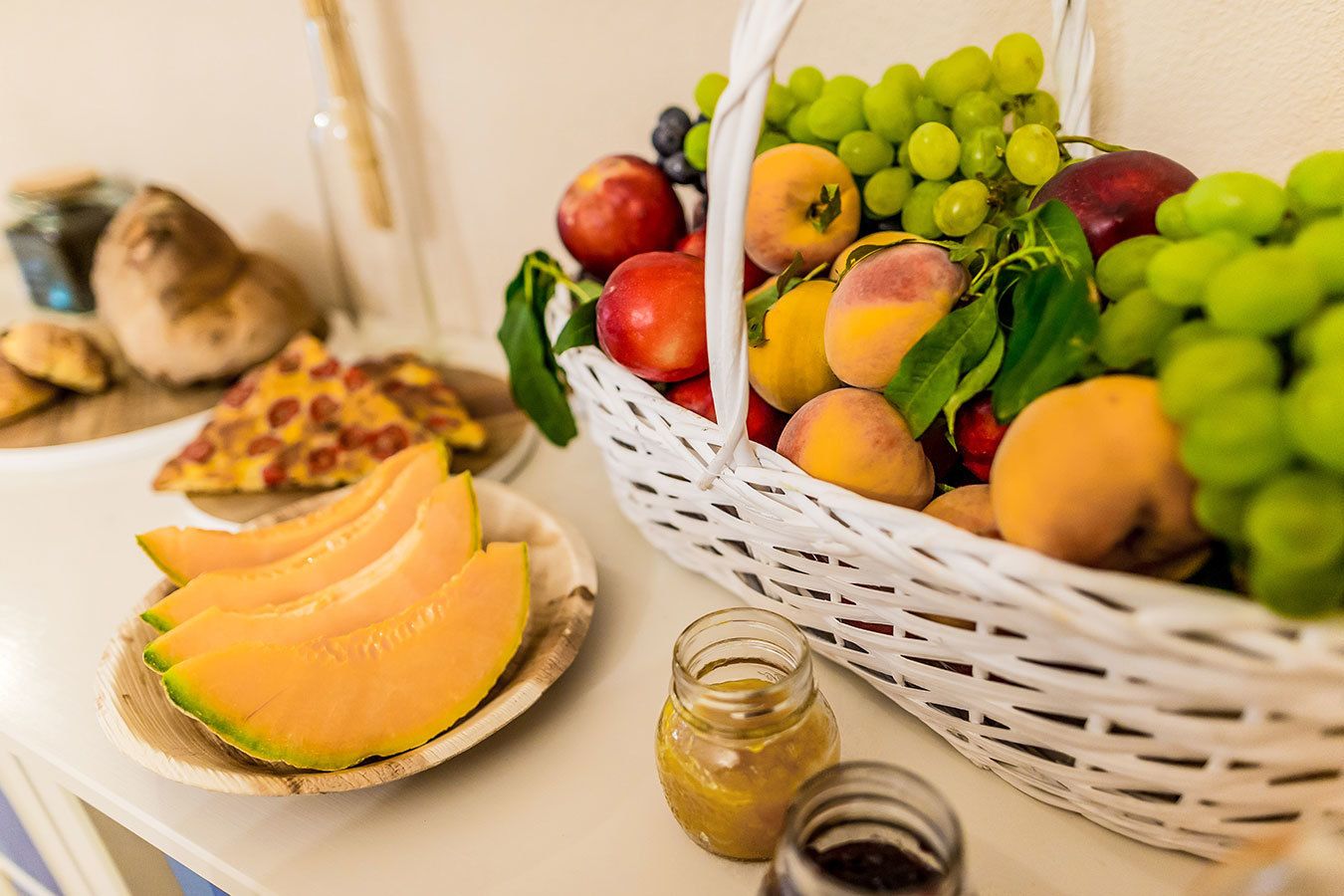 A table with a basket of fruits, a plate of melon slices, and two jars of preserves.