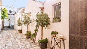 Un estrecho callejón empedrado con olivos en macetas y una mesa de madera con una cesta de flores.