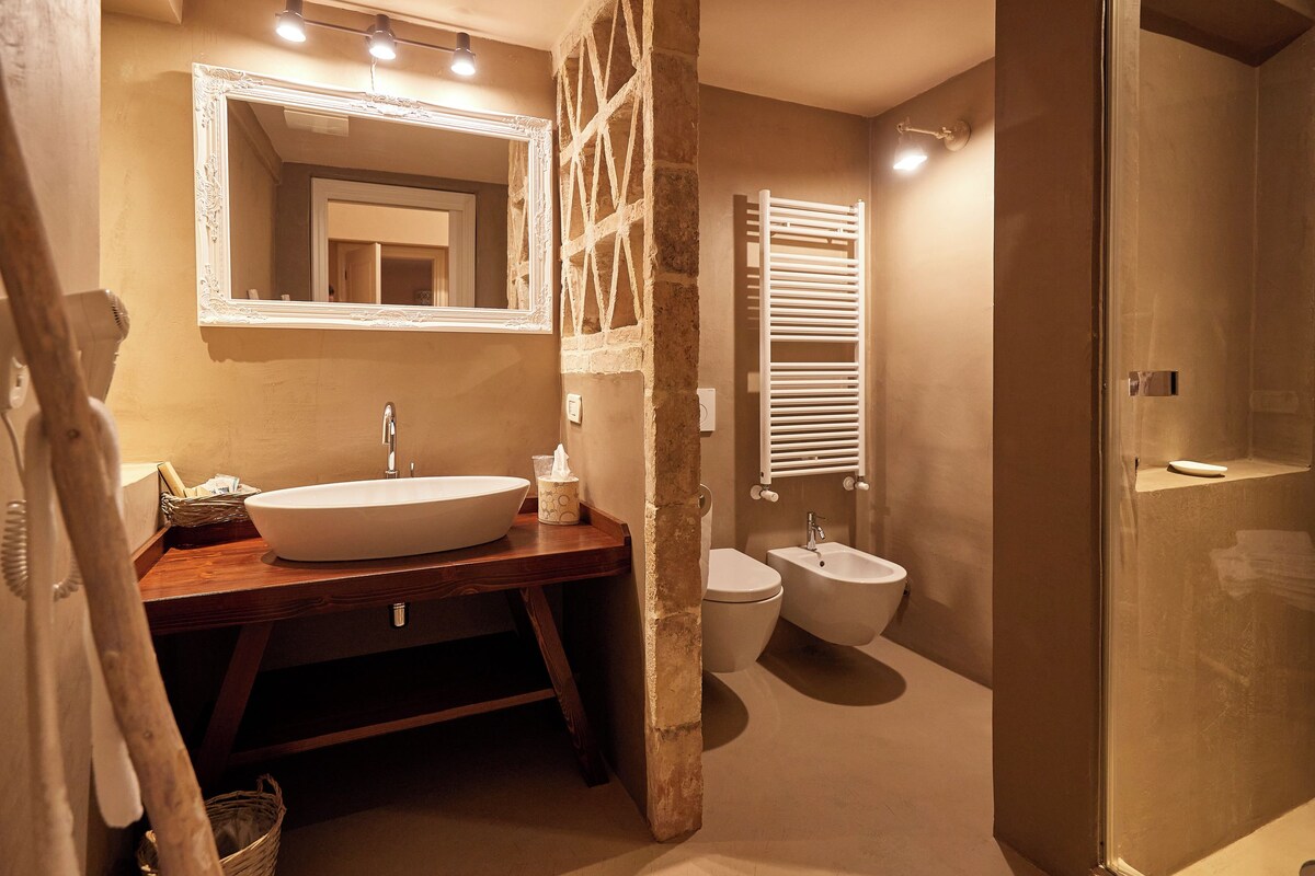 A bathroom with a large white sink, a wooden vanity, a freestanding mirror, a bidet, and a glass-enclosed shower.