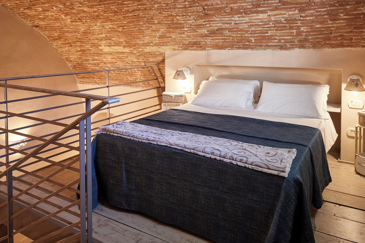 A bedroom with a brick wall, a bed with white linens, a dark blue bedspread, and a nightstand with a lamp.