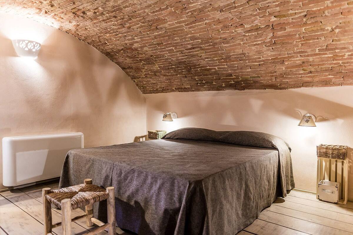 A cozy bedroom with a brick ceiling, a bed with a grey bedspread, two wall-mounted lamps, a wicker chair, and a radiator.