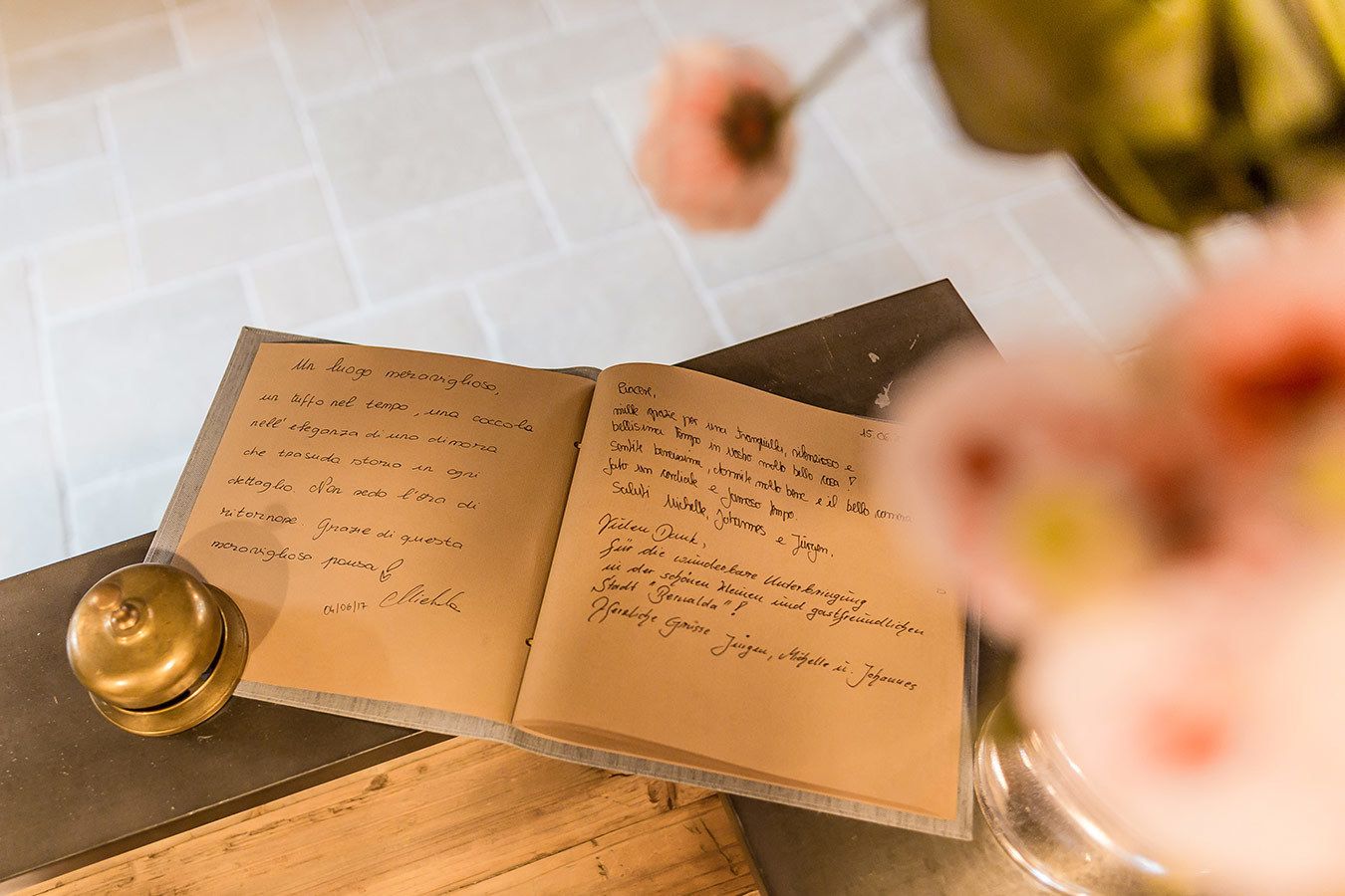 Ein offenes Buch mit handschriftlichem Text auf einer Holztischplatte, ein Messinggegenstand und eine Vase mit Blumen.