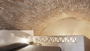 Una habitación con techo de ladrillo, una cama con cubrecama gris, una lámpara fijada a la pared y un panel decorativo en la pared.