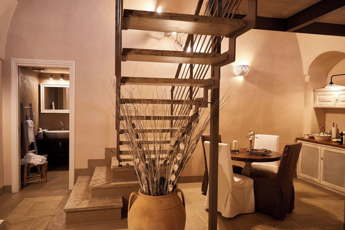 A staircase with a wooden railing leading to a second floor, a vase with dried plants in the foreground, a dining area with a table and chairs, and a kitchen area with cabinets and a sink.